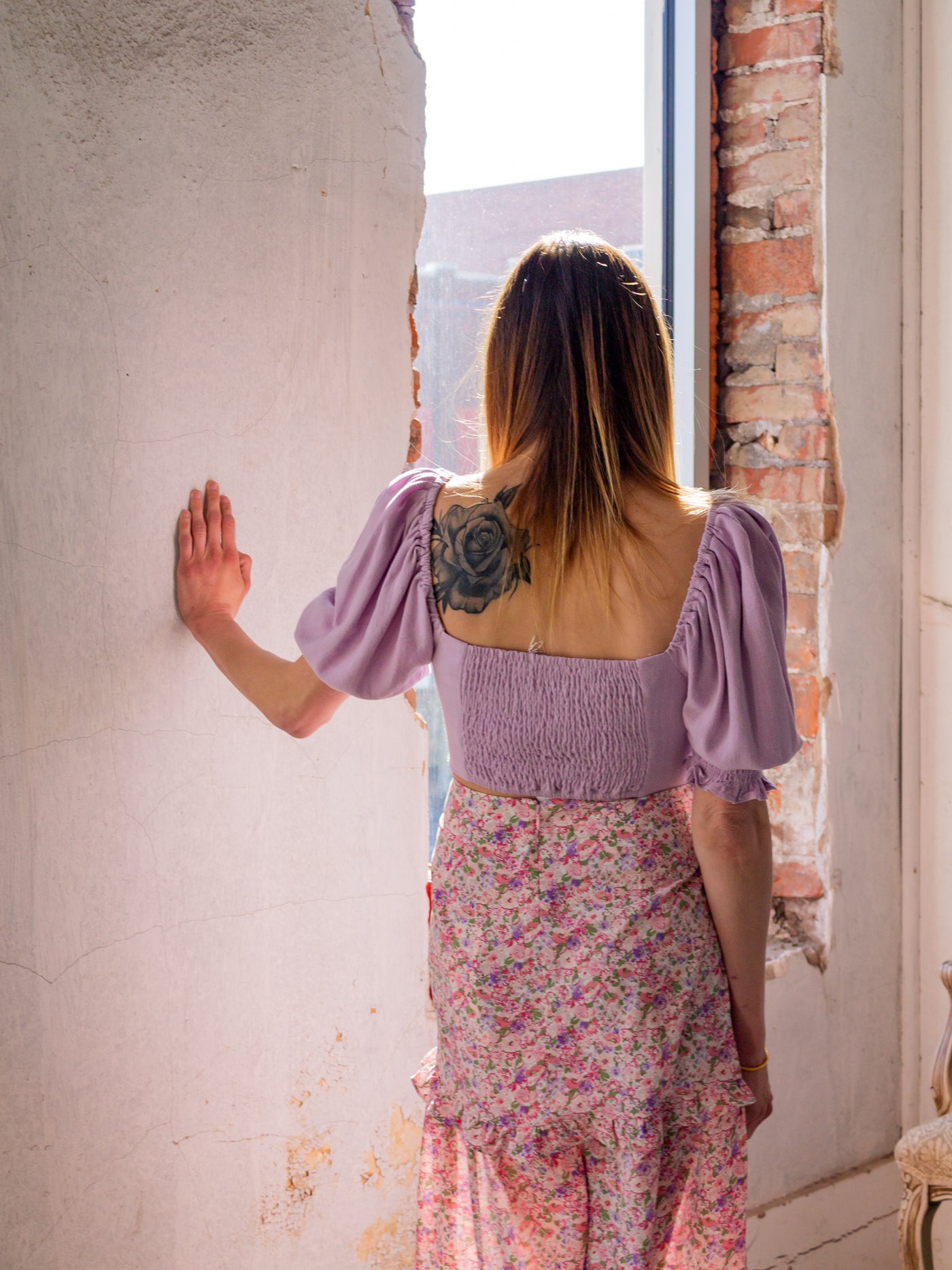 A model wearing a lavender crop top that has short sleeves with ruching, ruched back, and ties in the front like a bow. The model has it on with a floral midi skirt that is purple, pink, and white and has a slit on the side and a ruffle about halfway down.
