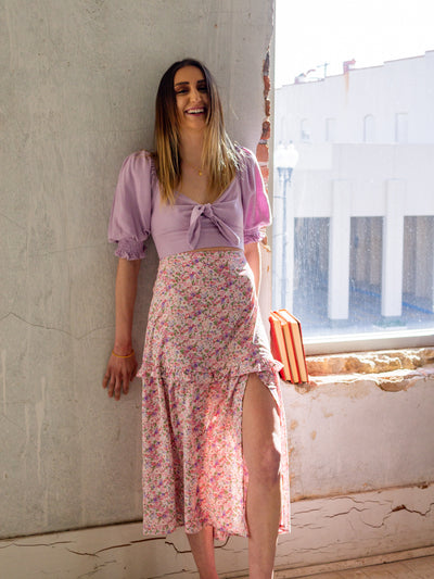 A model wearing a lavender crop top that has short sleeves with ruching, ruched back, and ties in the front like a bow. The model has it on with a floral midi skirt that is purple, pink, and white and has a slit on the side and a ruffle about halfway down.