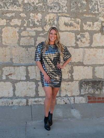 A model wearing a loose style mini dress with a black and silver sequin material. The dress has oversized balloon sleeves. The model has it on with black boots. 