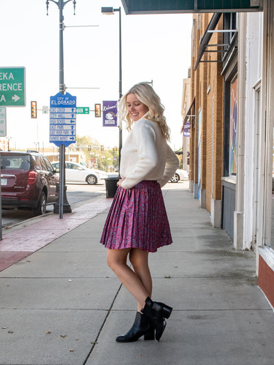 A model wearing a purple retro patterned pleated mini skirt. The model has it paired with a white turtleneck and black booties.