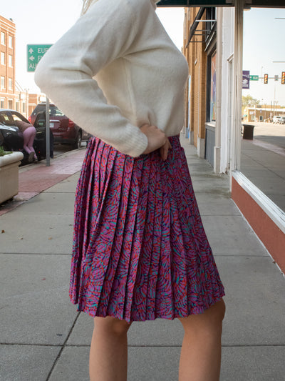 A model wearing a purple retro patterned pleated mini skirt. The model has it paired with a white turtleneck and black booties.