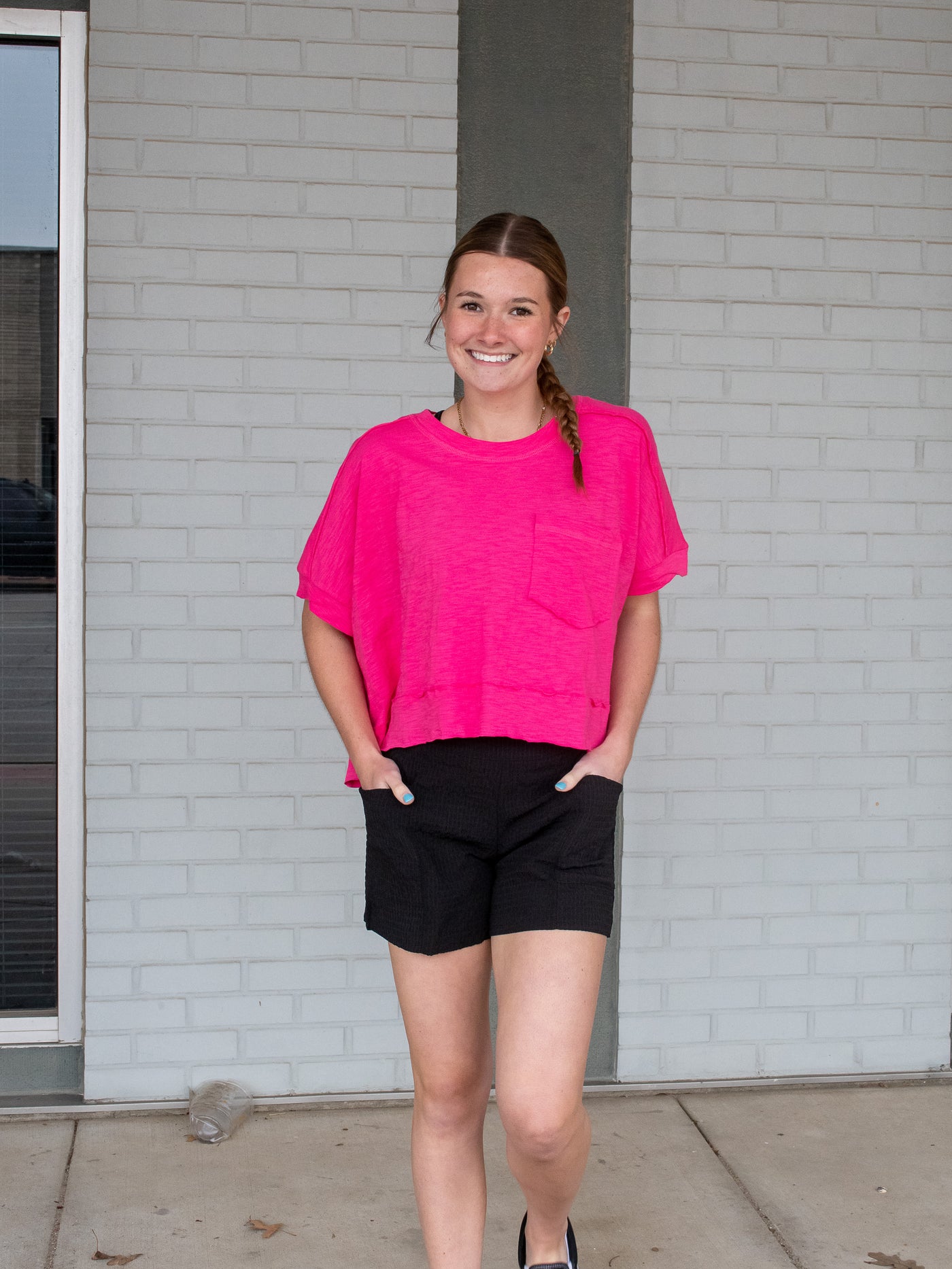 A model wearing a pink colored boxy slub jersey knit tee with raw edges and a front pocket. She has it on with black shorts and white sneakers.