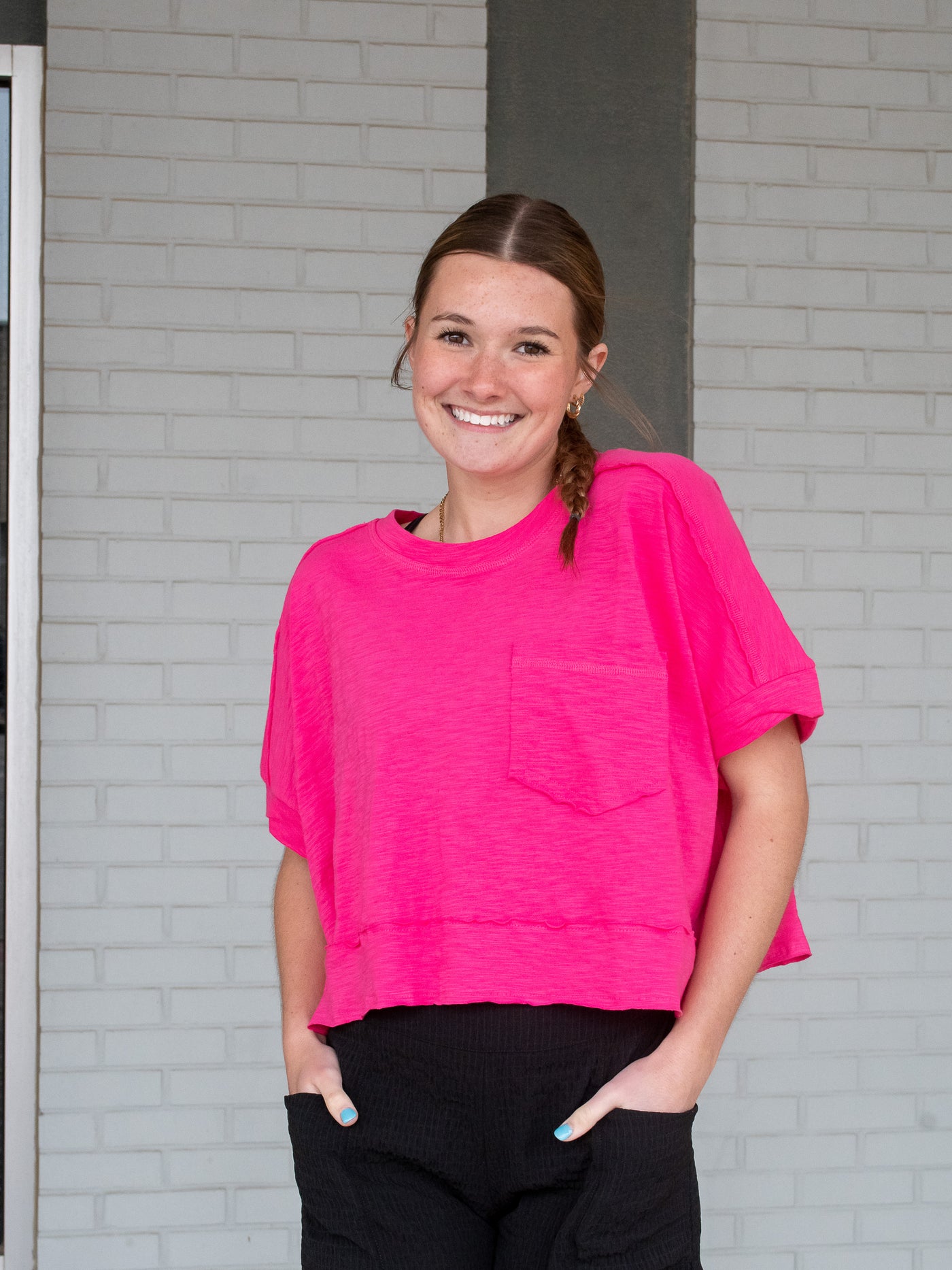 A model wearing a pink colored boxy slub jersey knit tee with raw edges and a front pocket. She has it on with black shorts and white sneakers.