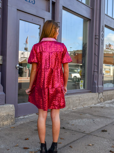 A model wearing a hot pink sequin dress with pouf sleeves that hits right above the knee. She has it on with black boots.