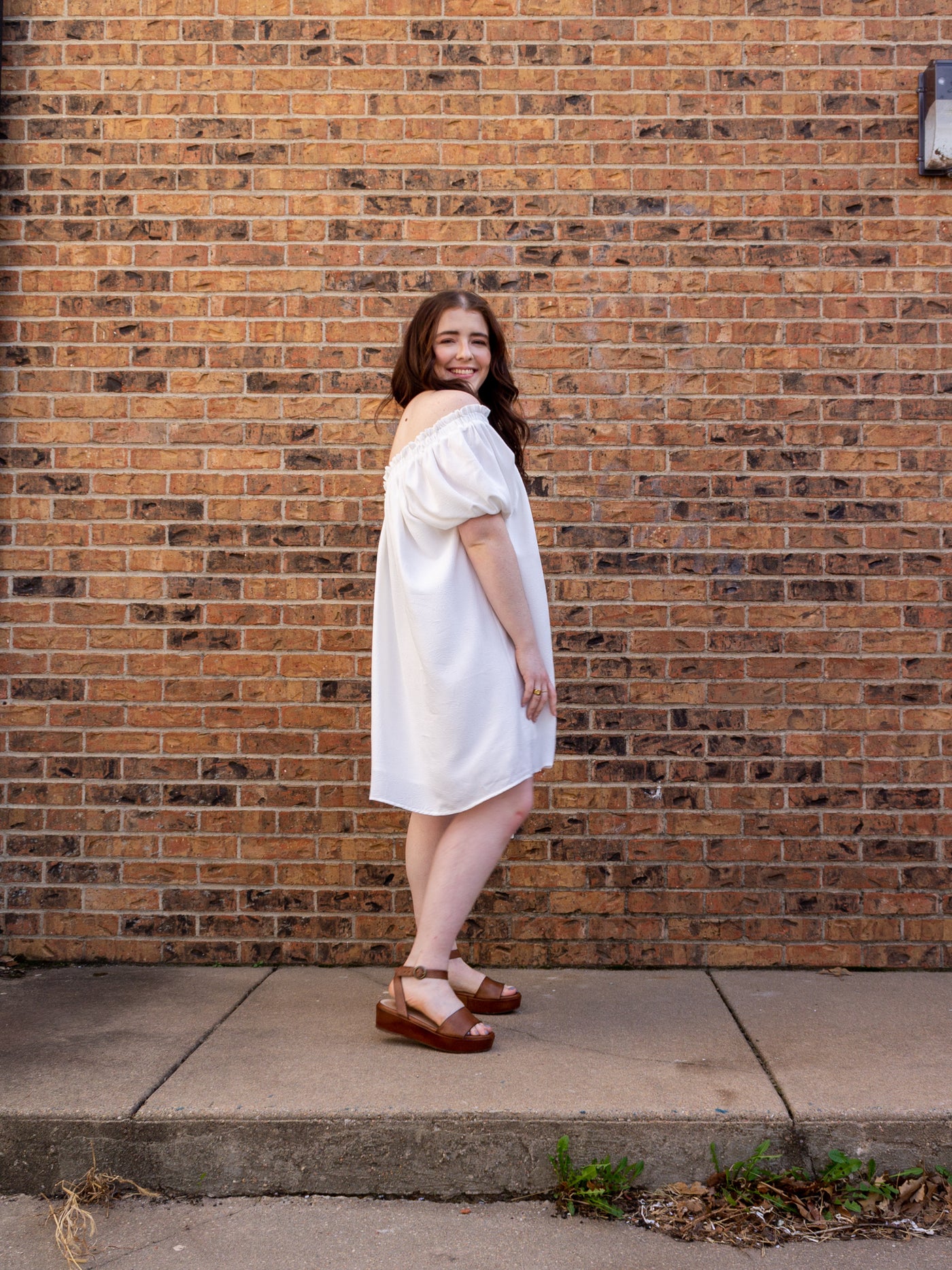 A model wearing a one shoulder, short sleeve white dress that hits right above the knee. The model has it on with a brown platform sandal.