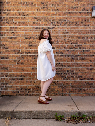 A model wearing a one shoulder, short sleeve white dress that hits right above the knee. The model has it on with a brown platform sandal.