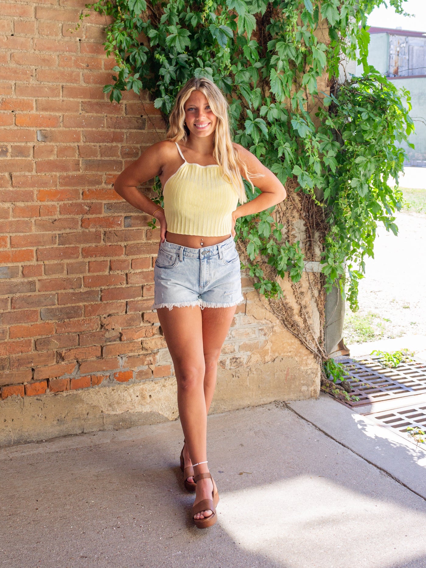 A model wearing a light yellow pointelle knit sleeveless crop top that ties in the back with a bow. She has it paired with denim shorts and a brown platform sandal.