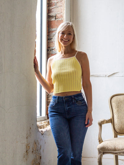A model wearing a light yellow pointelle knit sleeveless crop top that ties in the back with a bow. The model has it on with mid-rise dark wash flare jeans and a white sneaker.