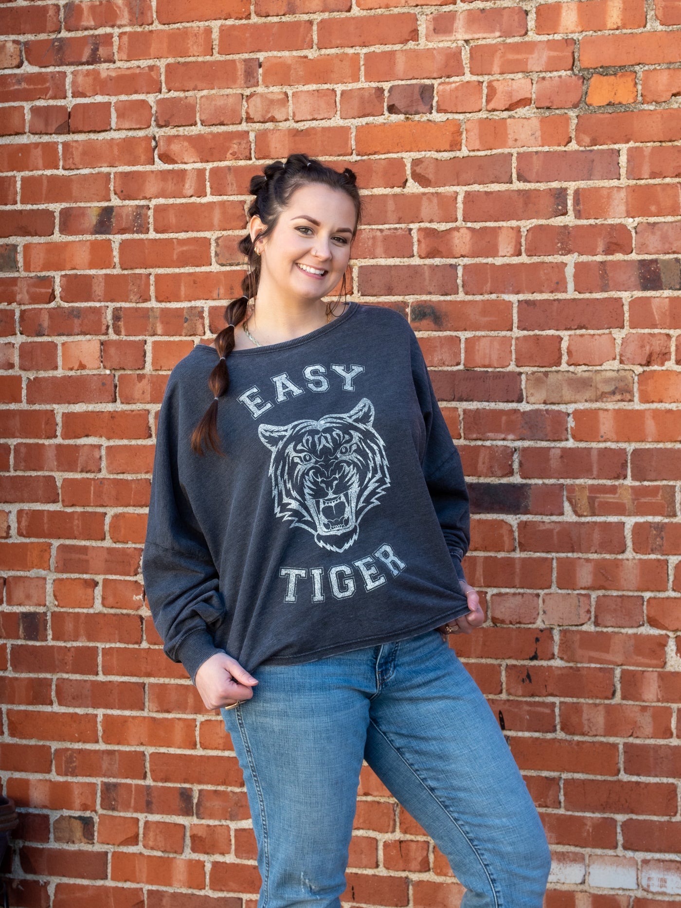 A model wearing a one size sweatshirt with an "Easy Tiger" graphic in a varsity font. It has a wide neck and high-low bottom. The model has it on with denim jeans.