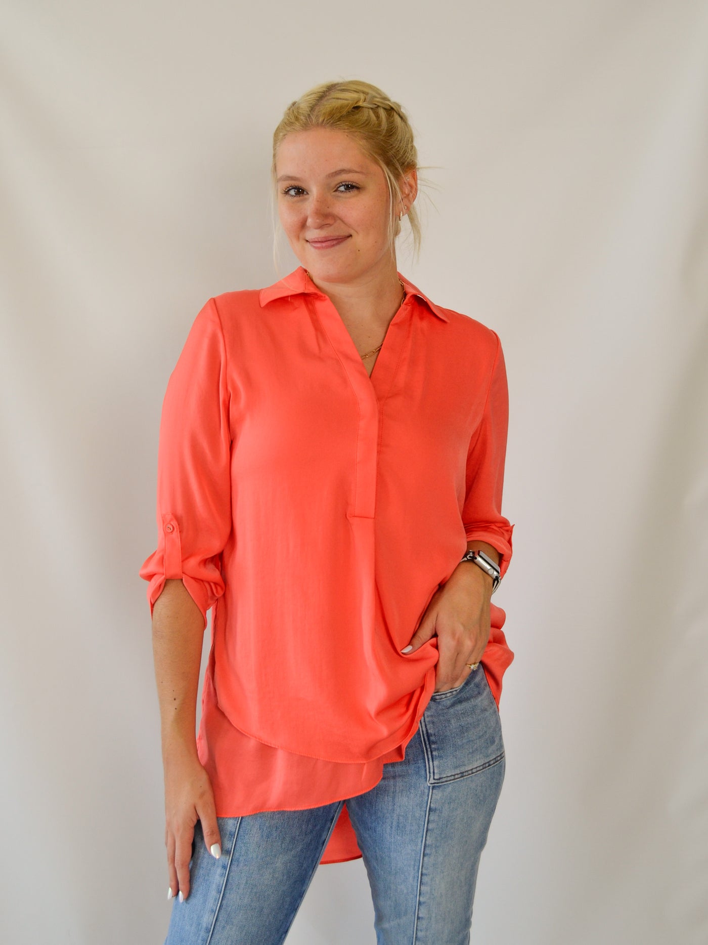 A model wearing a coral peach colored airflow blouse. The blouse is longer in the back than the front. She has it paired with denim jeans.