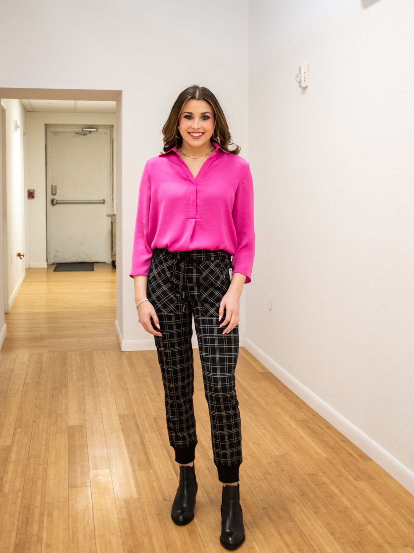 A model wearing a pink airflow blouse. The blouse is longer in the back than the front. She has it paired with plaid joggers.
