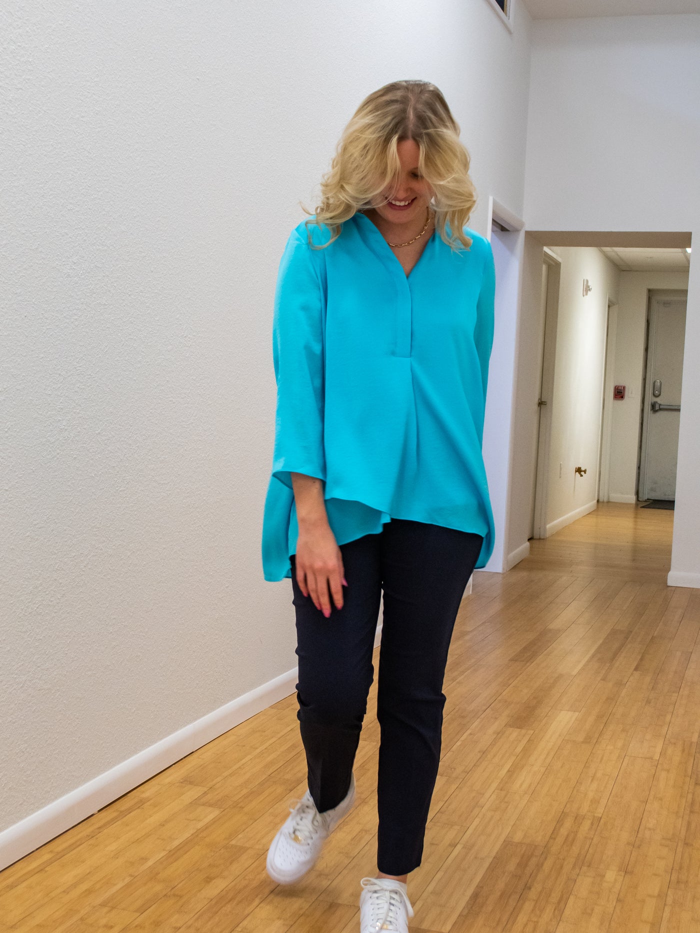 A model wearing a turquoise airflow blouse. The blouse is longer in the back than the front. She has it paired with navy dress pants.