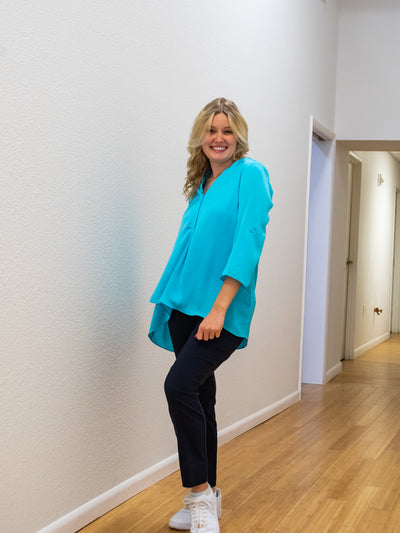 A model wearing a turquoise airflow blouse. The blouse is longer in the back than the front. She has it paired with navy dress pants.