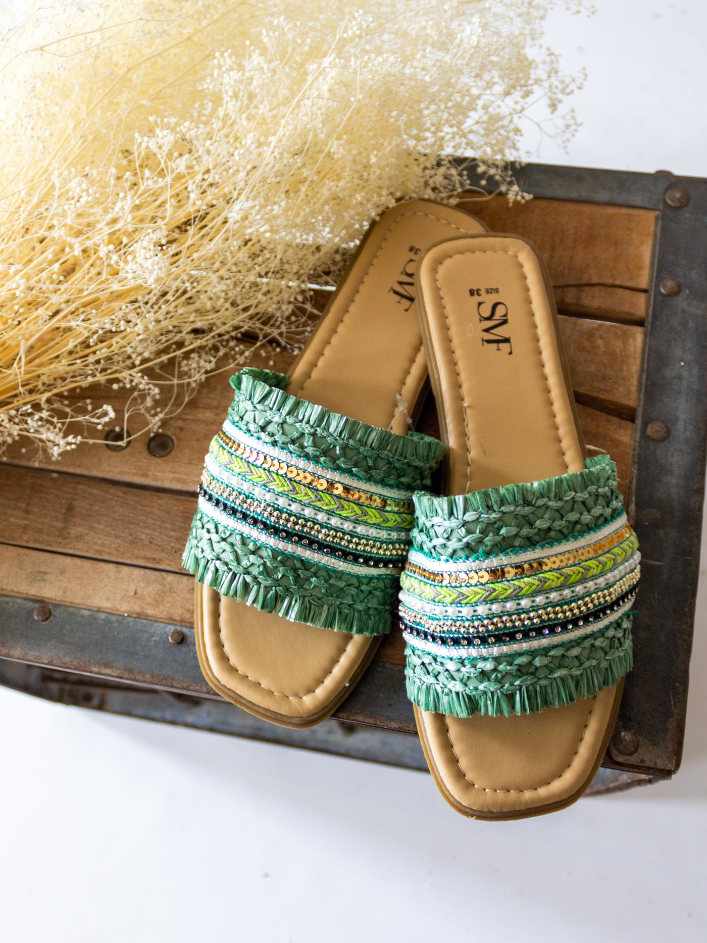 A boho themed sandal with braided, beads, sequins and other little accents on the top strap of the slide. They are shades of green, blue, gold, white, and black.