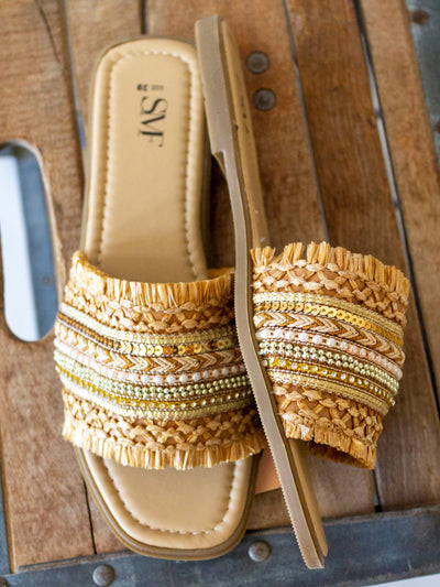 A boho themed sandal with braided, beads, sequins and other little accents on the top strap of the slide. They are shades of gold, brown, tan; neutral colors.