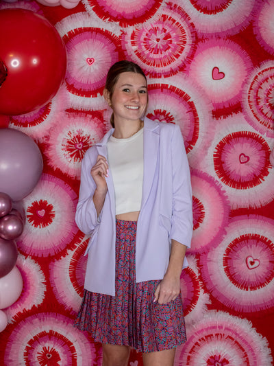 A model wearing a lilac shirred sleeve blazer over a white tank with a purple pleated skirt.