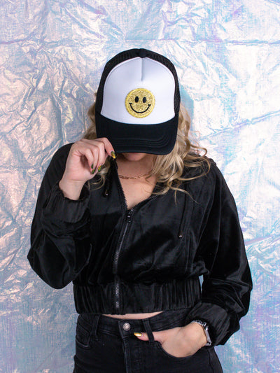 A model wearing a black trucker hat with a yellow smiley face on the front.