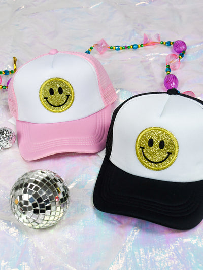 A pink trucker hat with a yellow smiley face patch on the front and a black trucker hat with a yellow smiley face on the front.