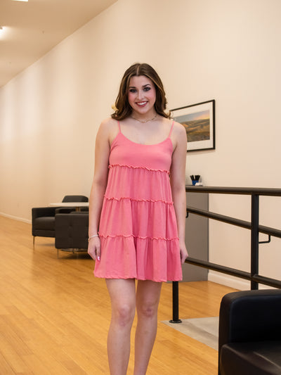 A model wearing a pink tiered mini dress with spaghetti straps and a scoop neck. She has it on with brown platform sandals.