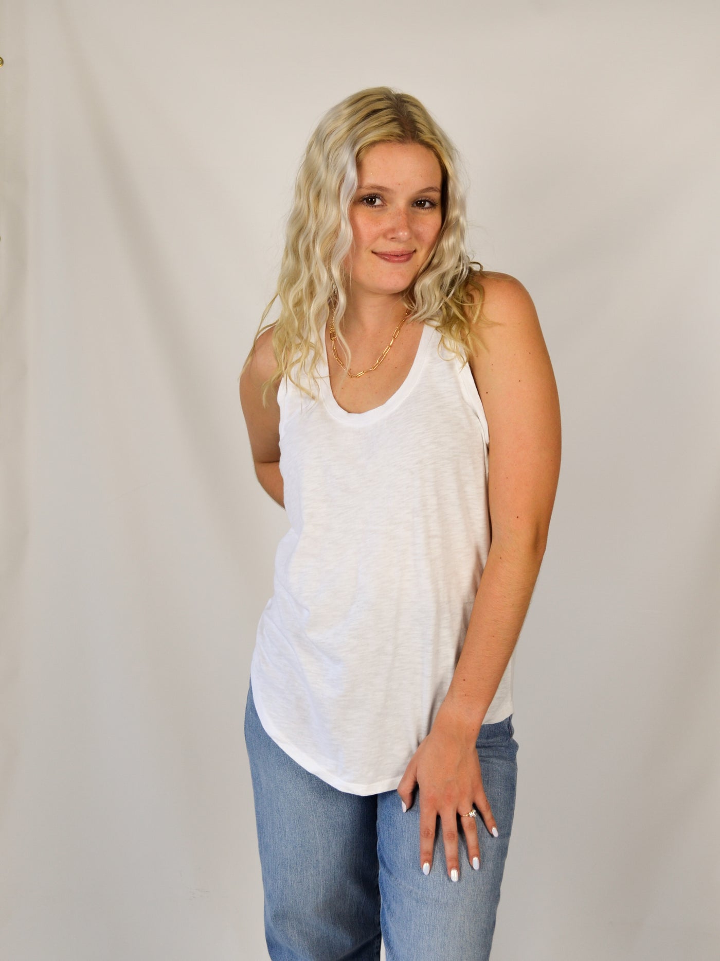 A model wearing a white slub, relaxed fit tank top with a curved hem. Th model has it on with light wash denim jeans.